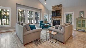 A spacious living room with two sofas, a fireplace, and large windows offering a view of trees.