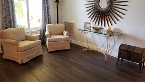 A living room with two beige armchairs, a glass coffee table with decorative items, a wall-mounted mirror, and a floor lamp.