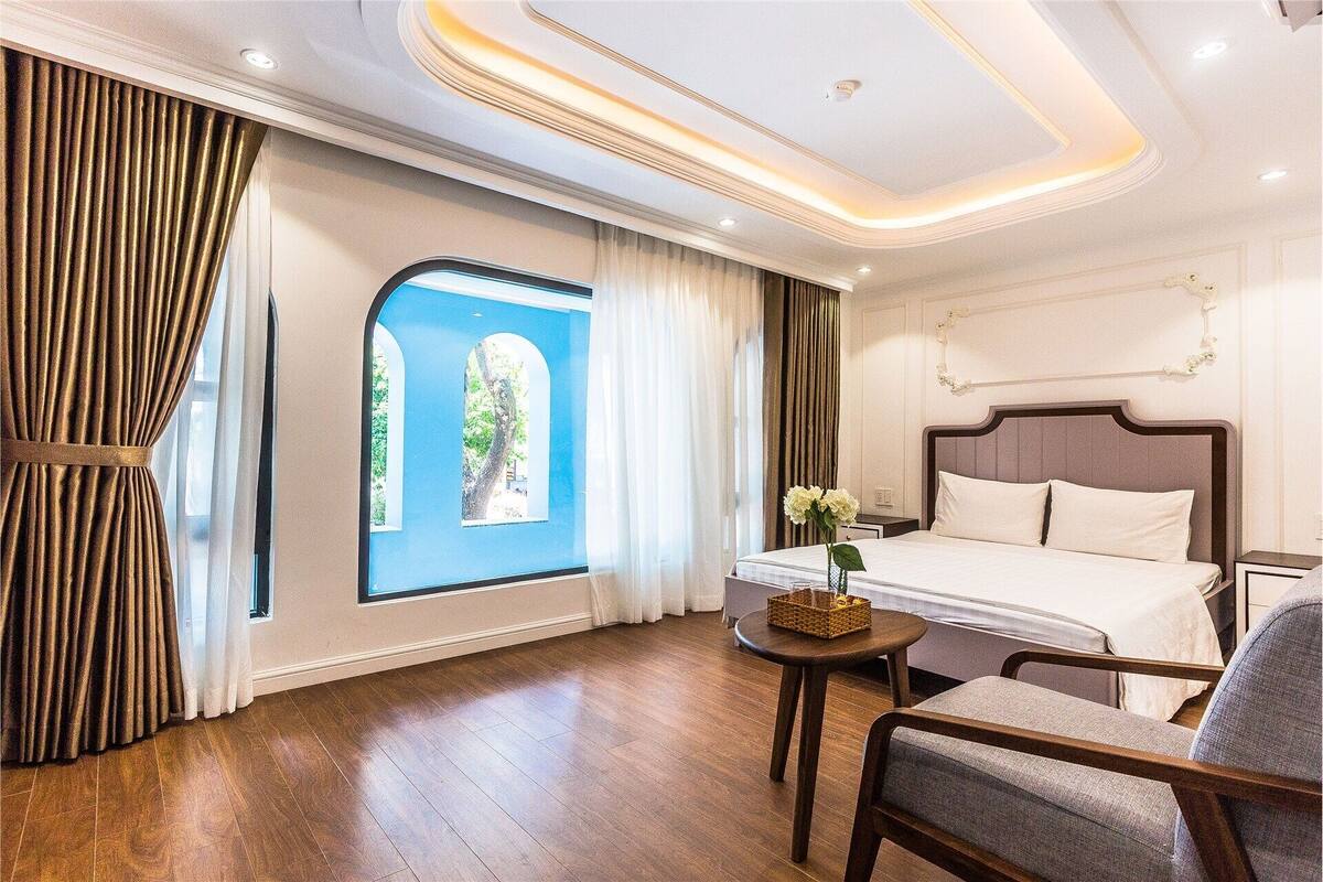 Habitación de hotel con cama, una silla, una mesita, ventana con cortinas y vistas a zonas verdes.