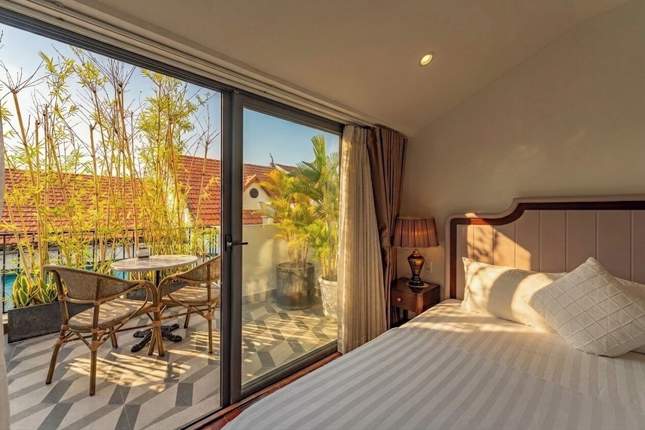A hotel room with a bed, a small table with chairs, a nightstand, and a view of a pool and palm trees.