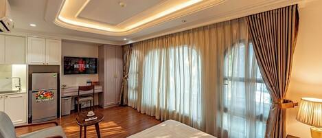 A hotel room with a bed, a desk with a TV, a kitchenette, and curtains.