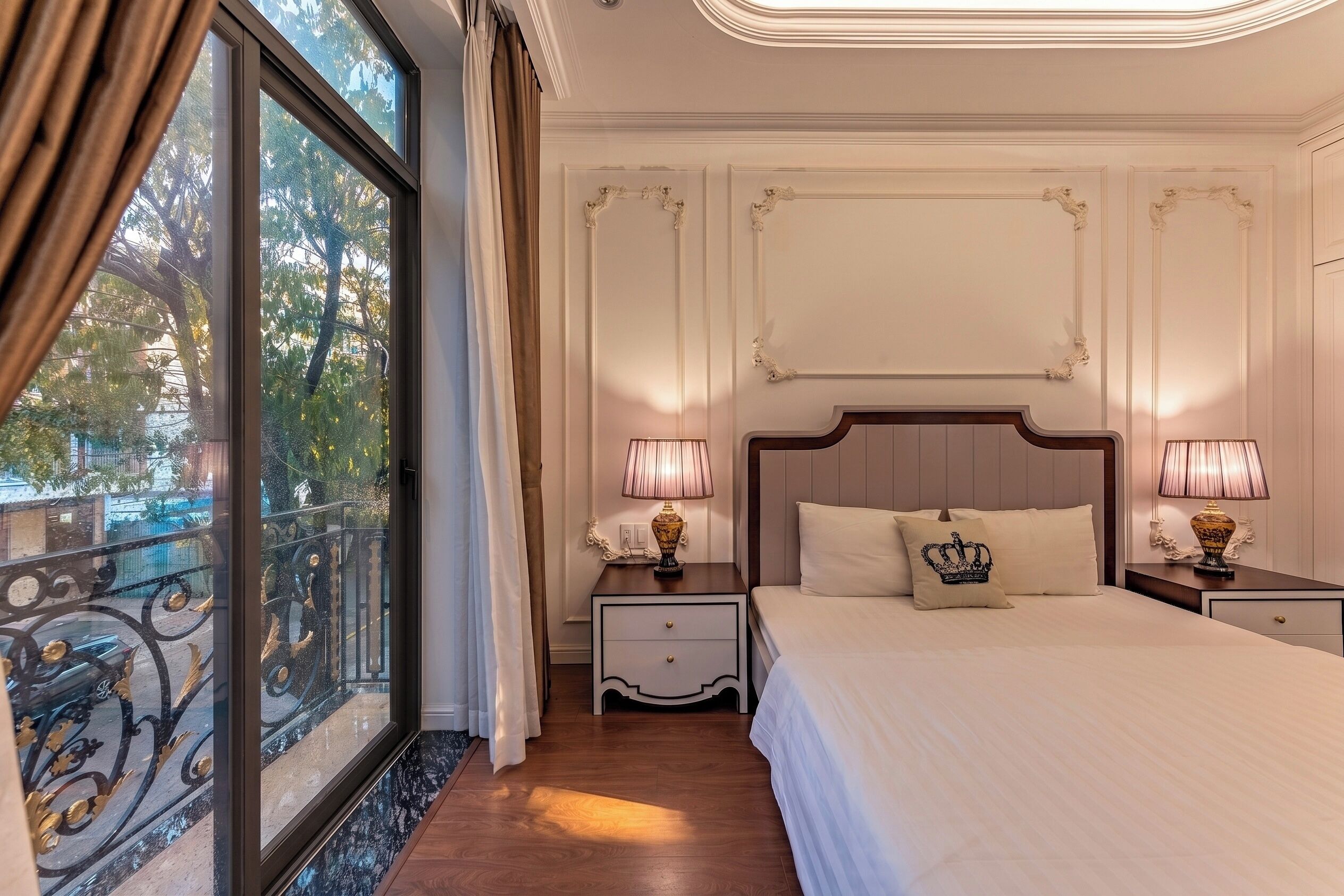 Habitación de hotel con una cama grande, dos mesitas de noche con lámparas, una mesita auxiliar, un ventanal con cortinas y un balcón con barandilla.