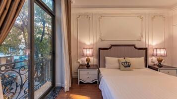 Habitación de hotel con una cama grande, dos mesitas de noche con lámparas, una mesita auxiliar, un ventanal con cortinas y un balcón con barandilla.
