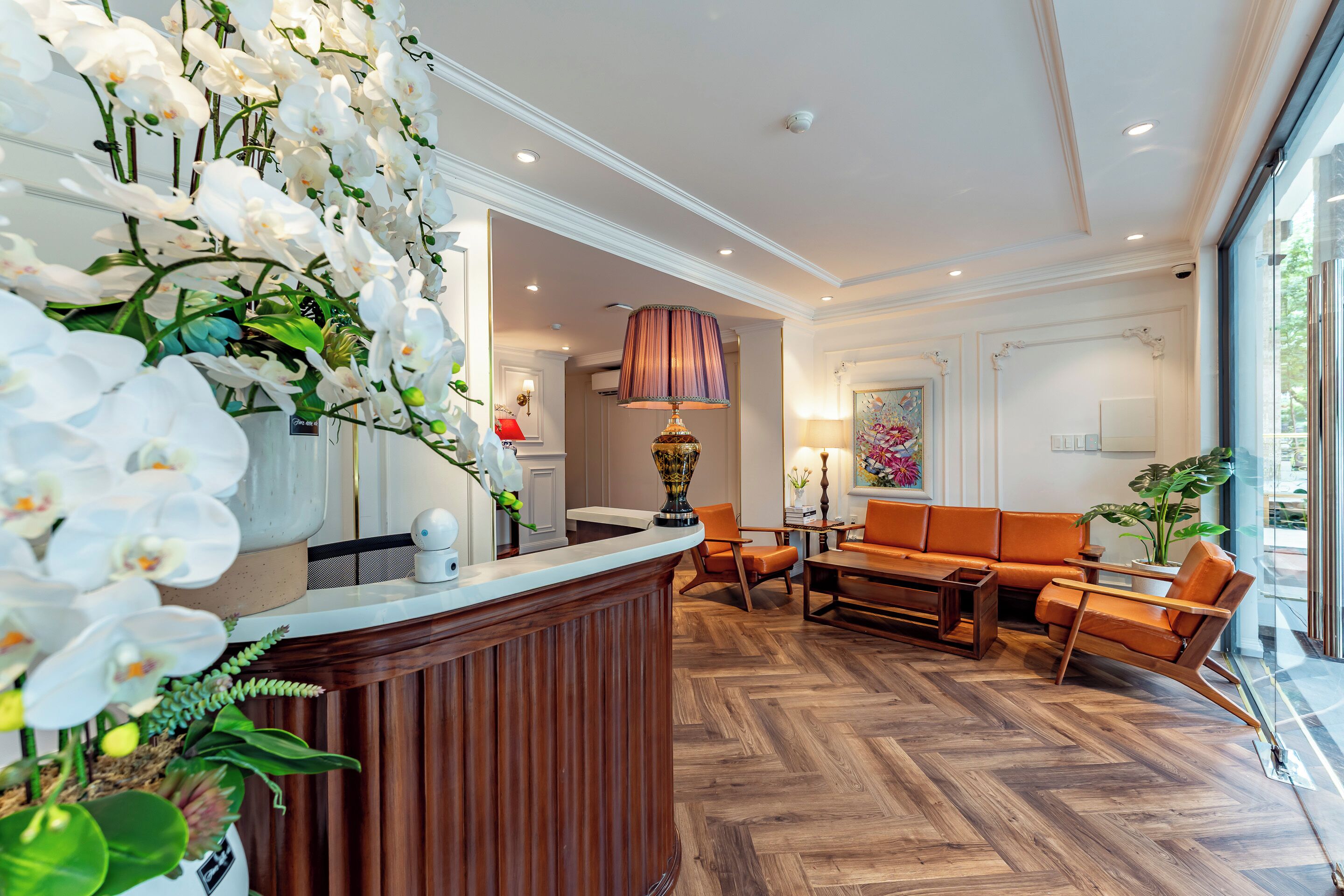 A reception area with a wooden reception desk, white orchids, a large lamp, a sofa, and a coffee table.