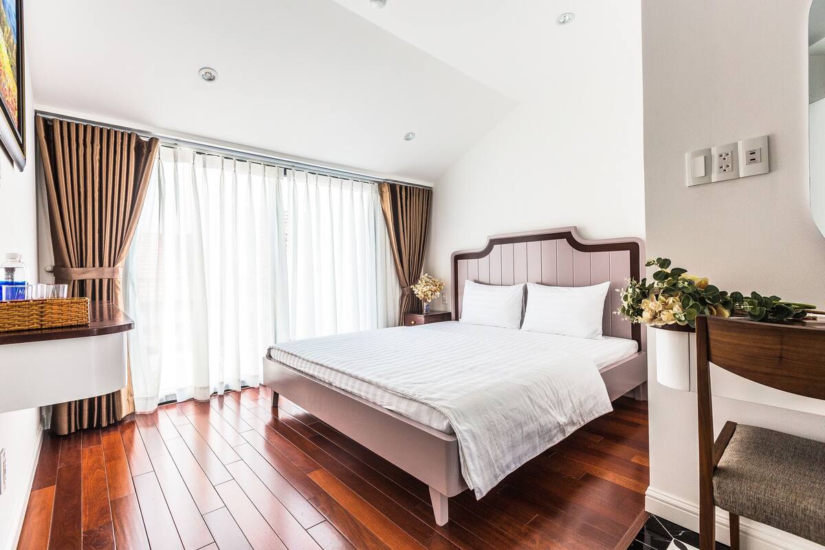 Un dormitorio con cama, mesita de noche, silla, ventana con cortinas y una lámpara de pared.