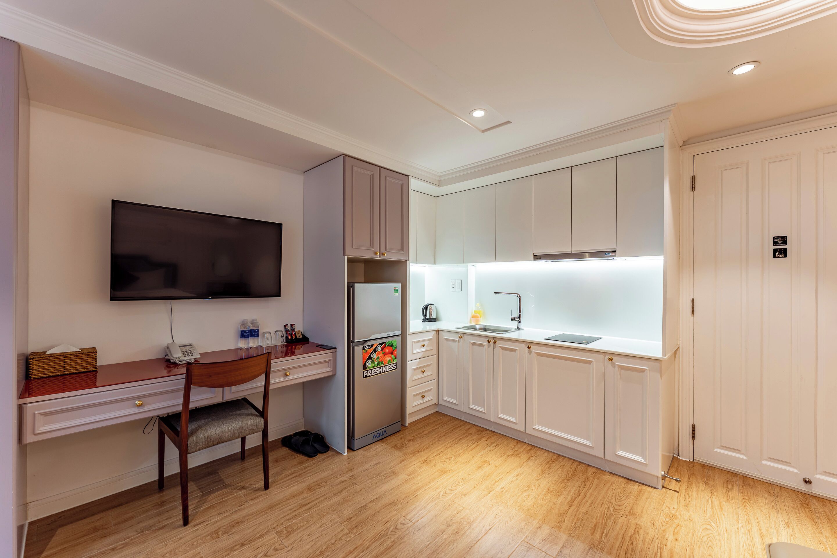 A compact kitchen with a wooden desk, a flat-screen TV, a refrigerator, and white cabinets.