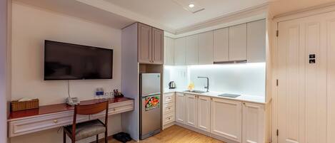 A compact kitchen with a wooden desk, a flat-screen TV, a refrigerator, and white cabinets.