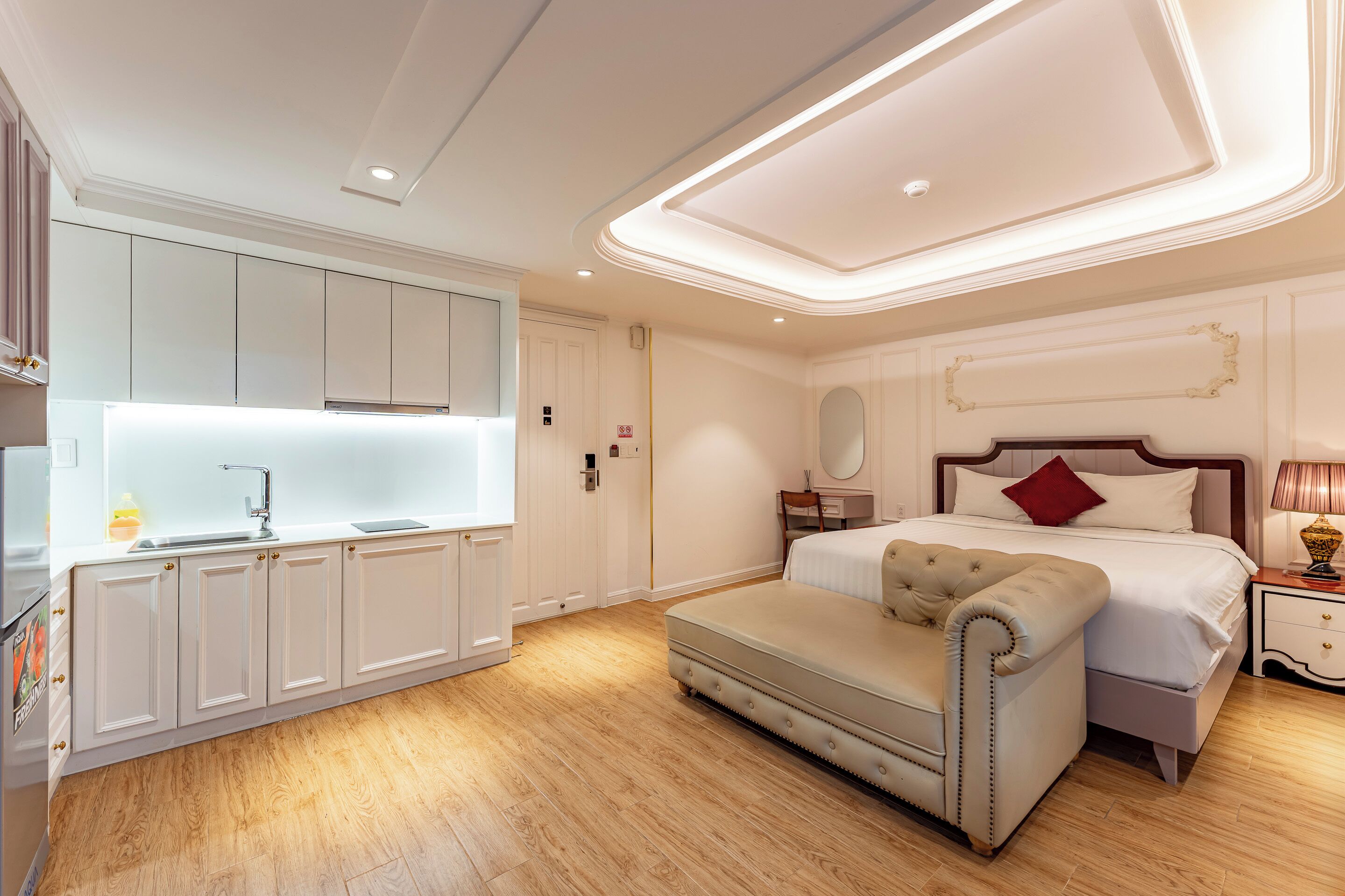 A modern bedroom with a large bed, a bedside table, and a nightstand. There is a small seating area with a cushioned bench. The room has wooden flooring and white walls.