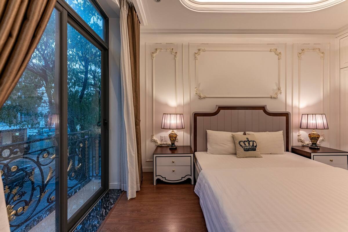 Habitación de hotel con una cama grande, dos mesitas de noche con lámparas, una mesita con lámpara, una ventana con cortinas y vista a árboles.