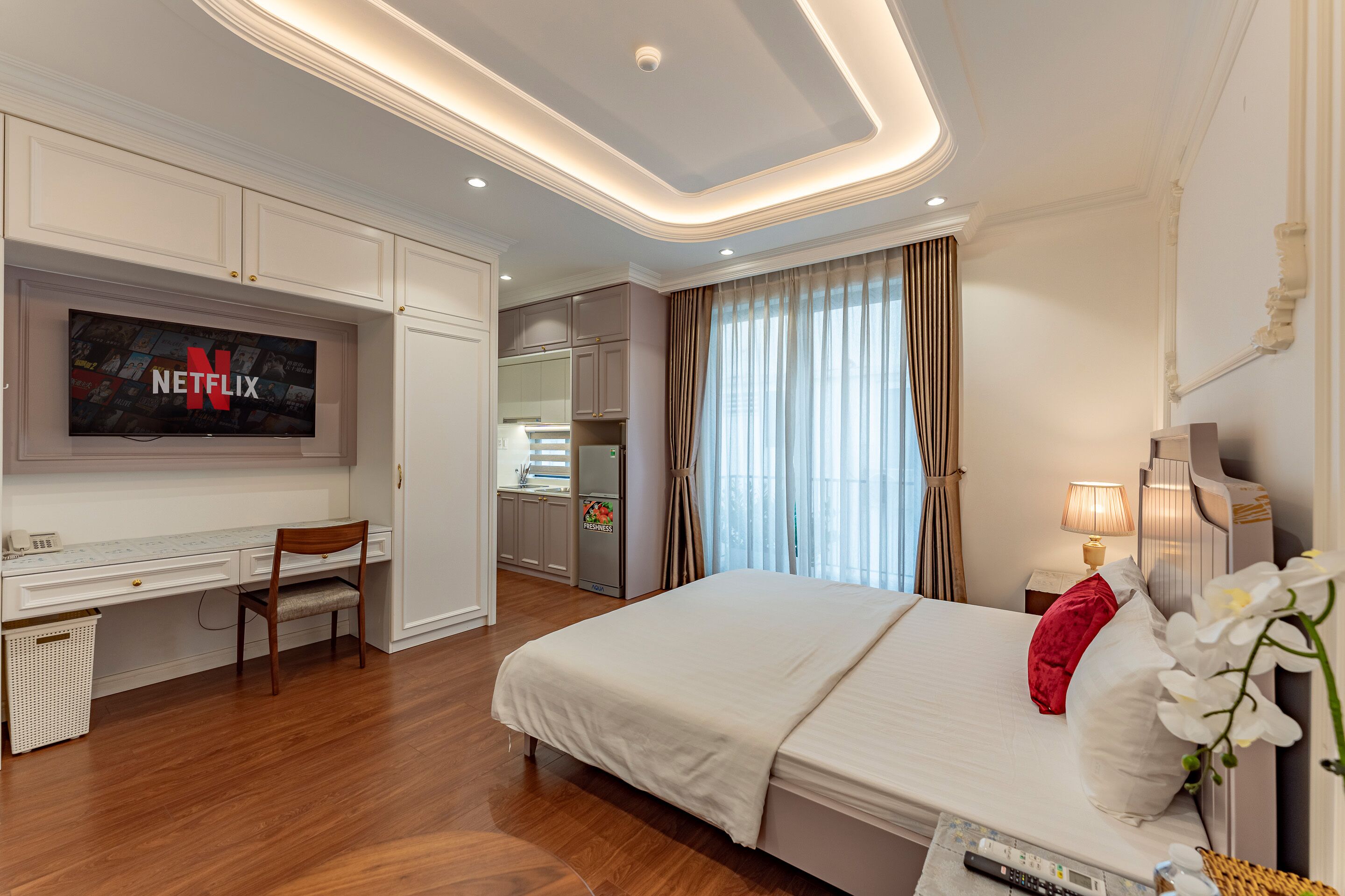 A modern hotel room with a large bed, a desk with a chair, a TV mounted on the wall, and a kitchenette in the background.