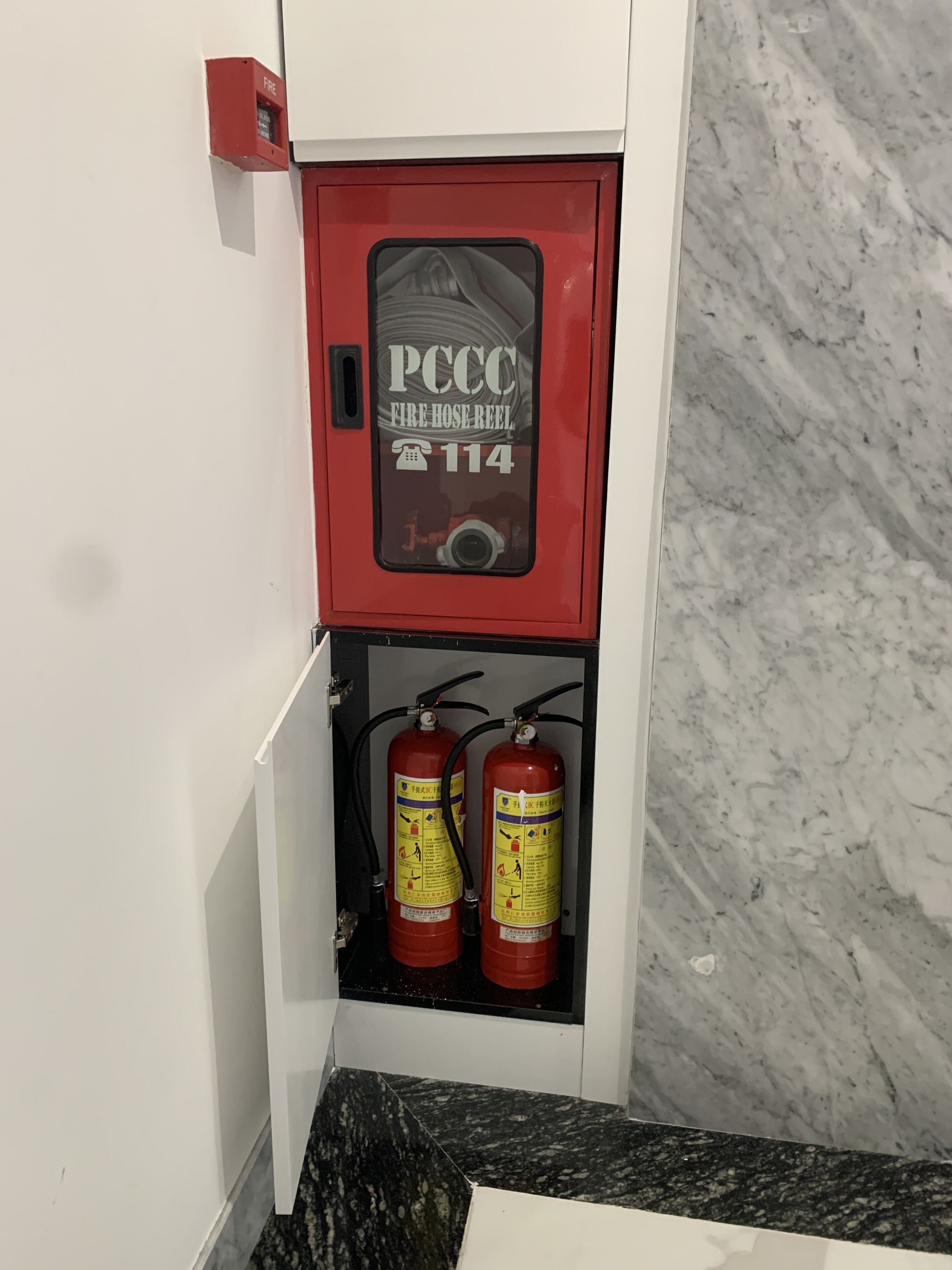 A fire hose reel cabinet with two fire extinguishers inside, a fire alarm, and a marble wall.
