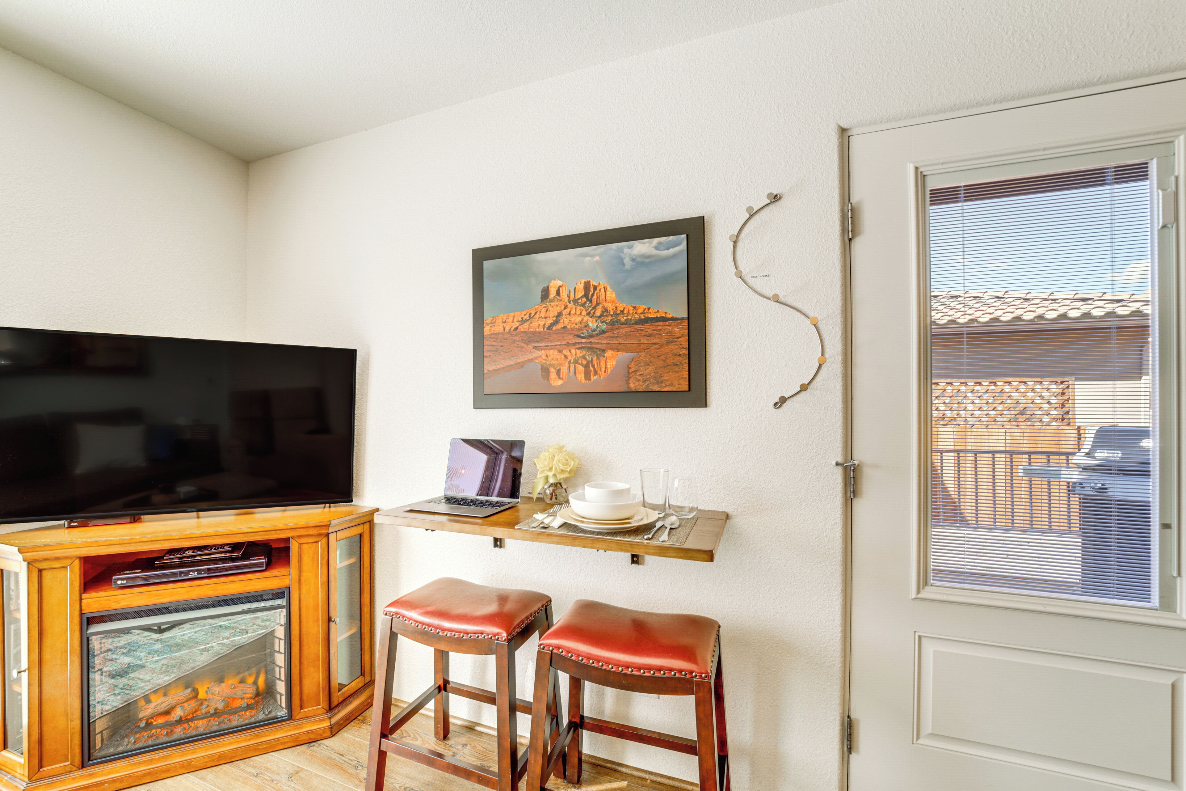 A living room with a flat-screen TV, a fireplace, two leather bar stools, a small wooden table with a laptop, a framed picture, and a window with blinds.