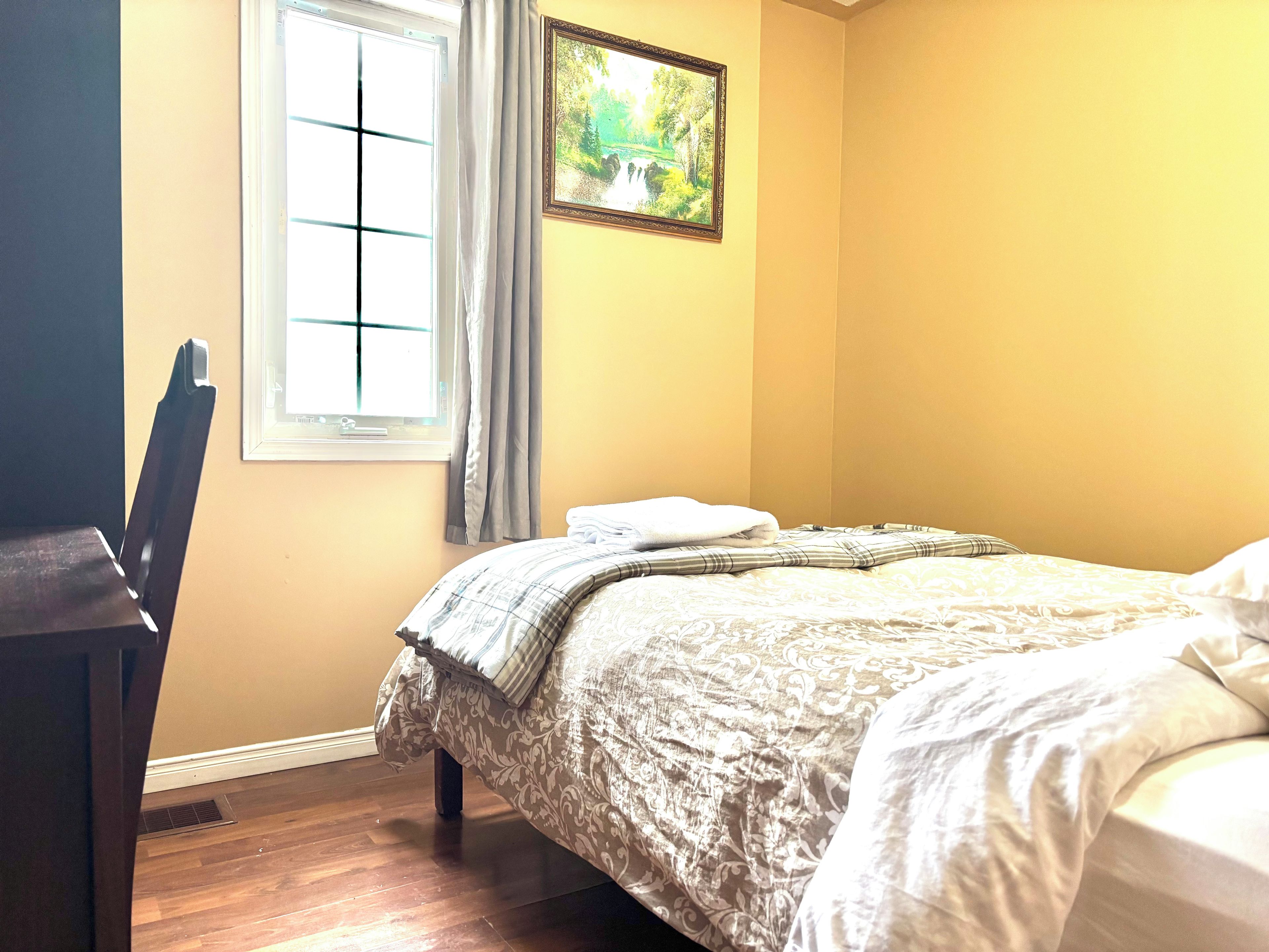 A bedroom with a bed, a chair, a window with curtains, and a framed picture on the wall.