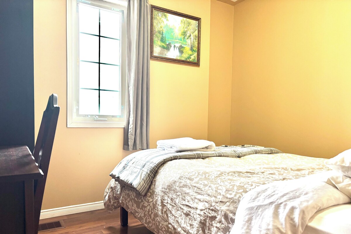 A bedroom with a bed, a chair, a window with curtains, and a framed picture on the wall.
