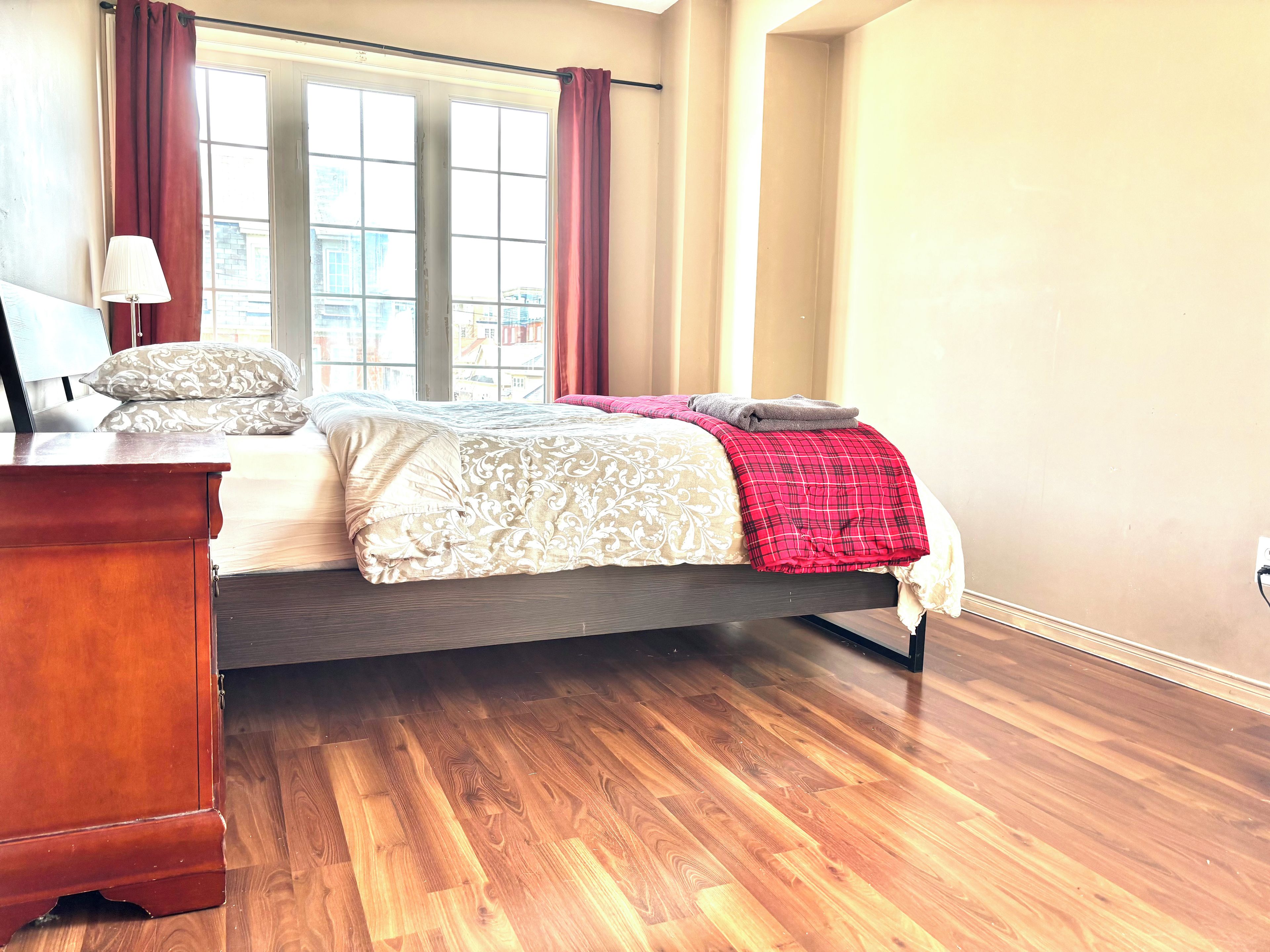 A bedroom with a bed, a wooden dresser, a window with curtains, and a lamp.
