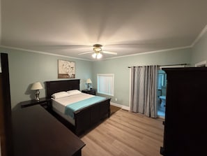 A bedroom with a bed, two bedside tables with lamps, a ceiling fan, a window with blinds, and a door leading to another room.