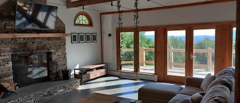 A spacious living room with a stone fireplace, wooden beams, a large sofa, and a view through large windows.