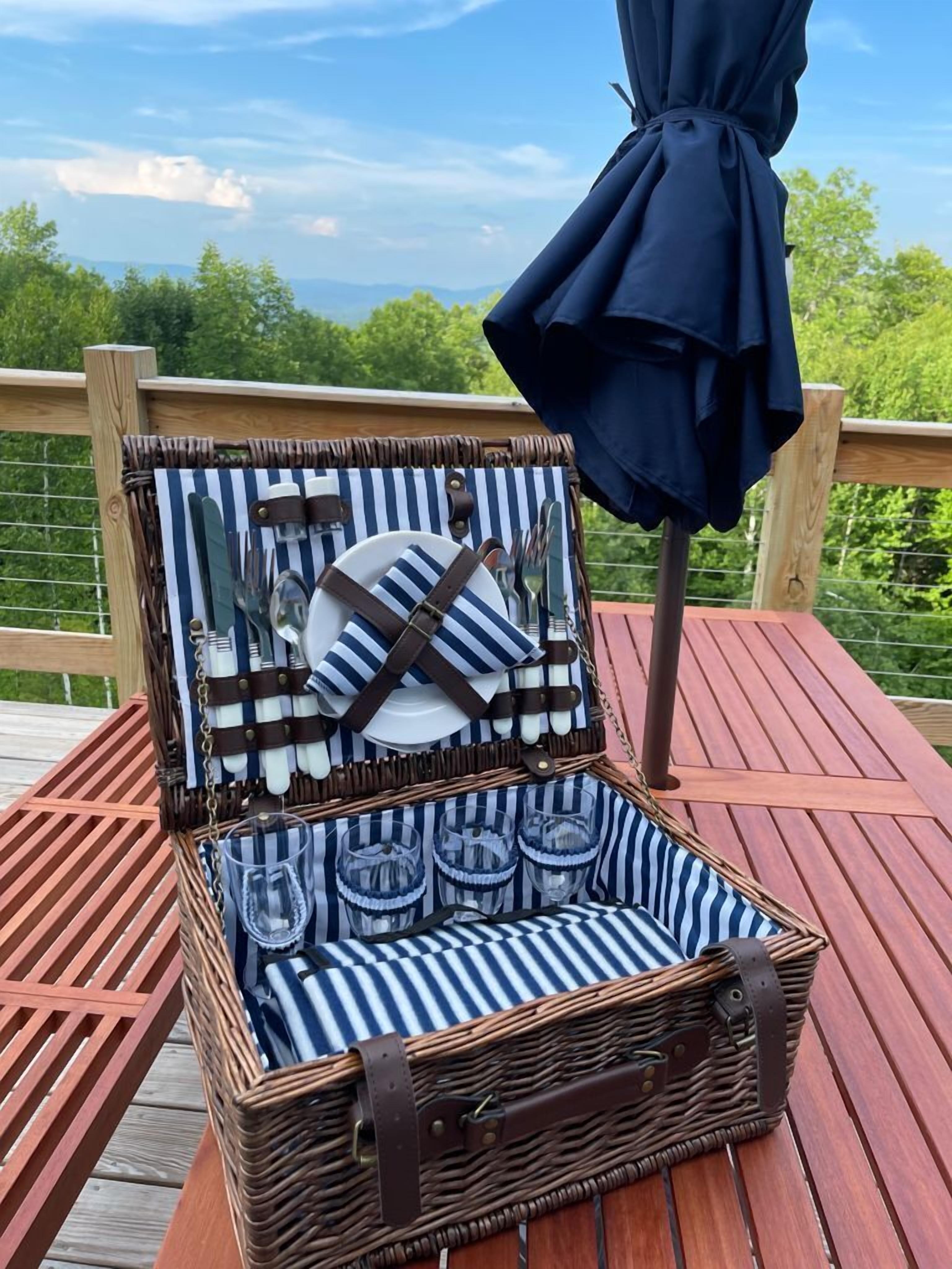 Un panier de pique-nique en osier ouvert, avec un intérieur à rayures, contenant des assiettes, des couverts et des verres, posé sur une terrasse en bois offrant une vue sur les arbres et un ciel dégagé.