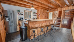 Interior de madera con una zona de cocina, taburetes de bar y un refrigerador de acero inoxidable.