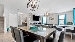 A modern living room with a glass-top dining table, black leather chairs, a pool table, and a flat-screen TV mounted on a wooden wall. - Villa 9020 - Near Parks, Free Pool/Spa Heat, Game Rm, & PS5! UPDATED MAY 2026! (Champions Gate)