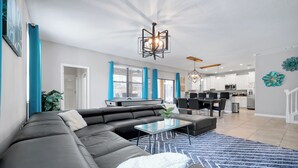 A modern living room with a sectional sofa, a glass coffee table, and a dining area with a kitchen in the background. - Villa 9020 - Near Parks, Free Pool/Spa Heat, Game Rm, & PS5! UPDATED MAY 2026! (Champions Gate)