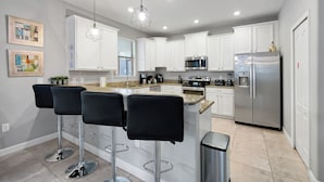 A modern kitchen with white cabinets, stainless steel appliances, and a central island with bar stools. - Villa 9020 - Near Parks, Free Pool/Spa Heat, Game Rm, & PS5! UPDATED MAY 2026! (Champions Gate)