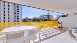 Een balkon met een geel veiligheidshek, een witte tafel en stoelen, en uitzicht op omliggende gebouwen.