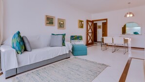 Een moderne woonkamer met een bank, een salontafel en een eethoek met stoelen. Er hangen fotolijstjes aan de muur en een deur die naar een andere kamer leidt.