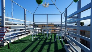 Terraza en la azotea con barandillas de metal blanco, césped artificial verde y sillas de metal blanco.