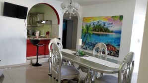 A dining area with a table and chairs, a painting of a tropical beach, and a kitchen in the background.
