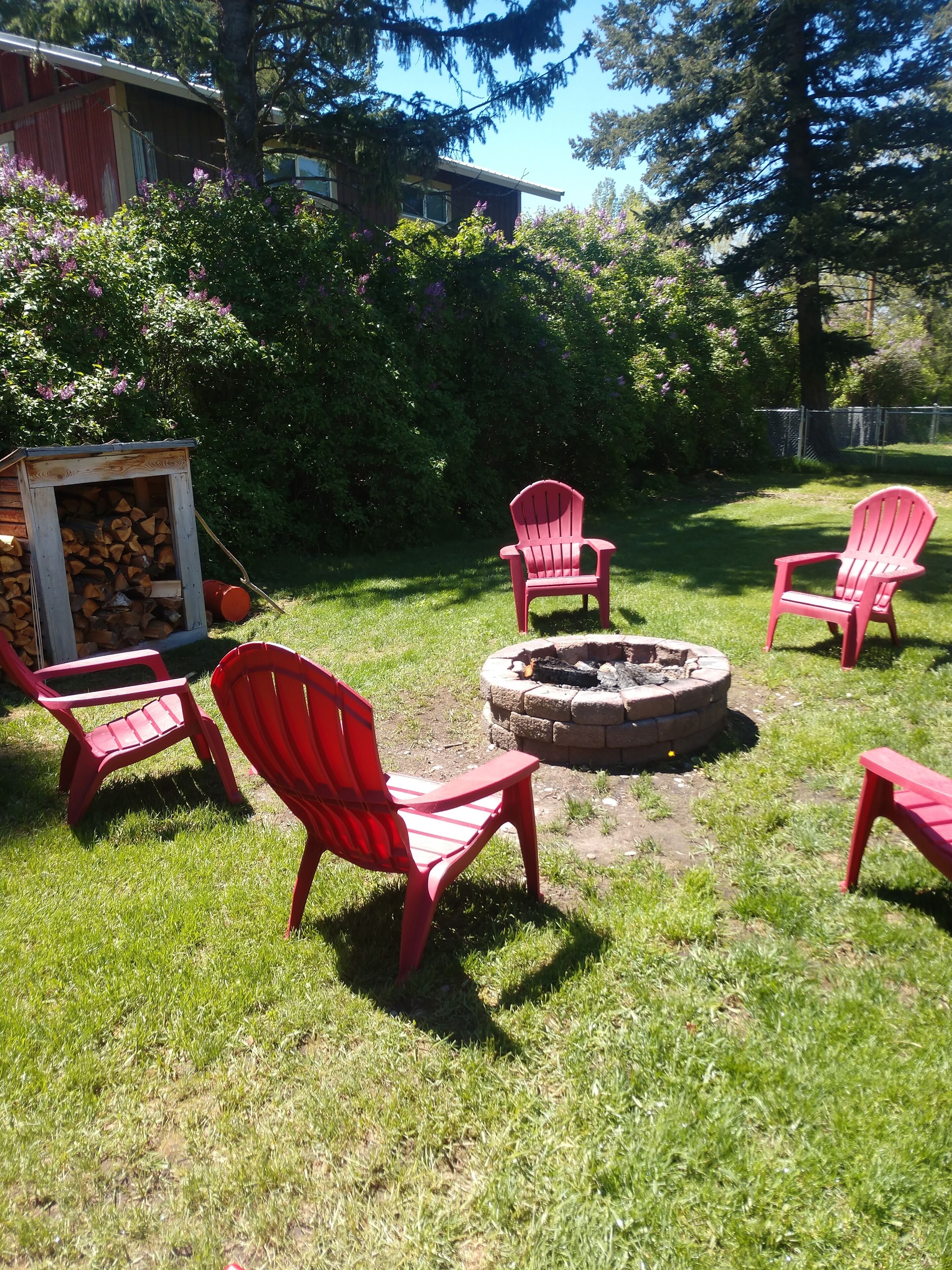 Un patio trasero con un brasero, sillas Adirondack rojas y una pila de leña.