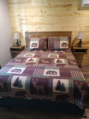 A bed with a quilt featuring animal and house patterns, flanked by two bedside tables with lamps, in a room with wooden walls. - Beautiful cabin close to Glacier Park, Flathead Lake & Whitefish Mountain. (Columbia Falls)