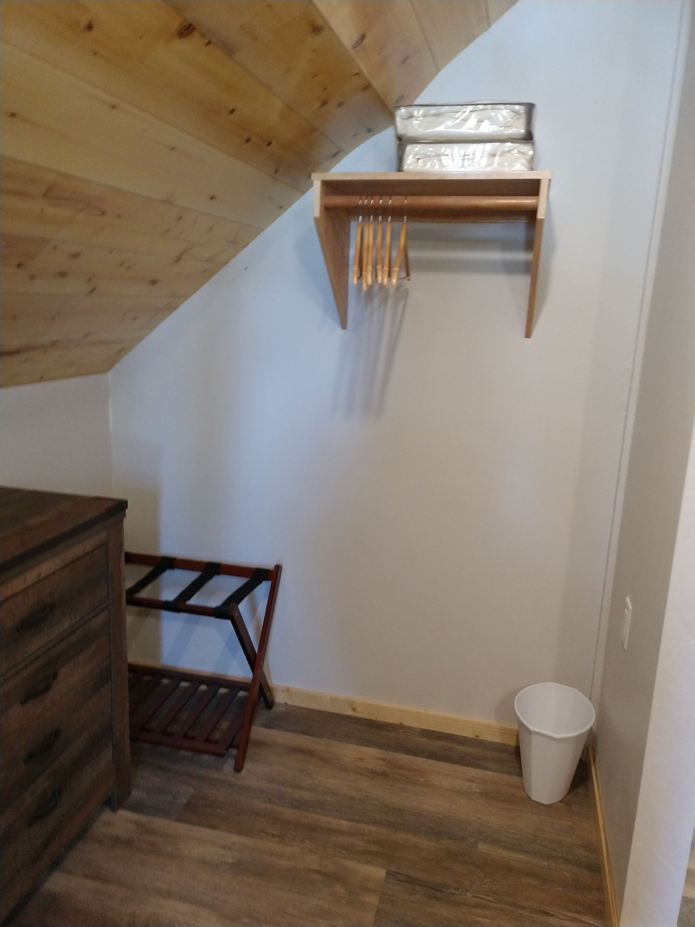 A room with a sloped ceiling, a wooden dresser, a metal rack with utensils, a wooden chair, and a white trash bin.