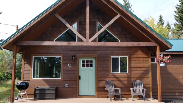 Une cabane en bois avec une porte verte, deux fenêtres et une véranda avec des chaises.