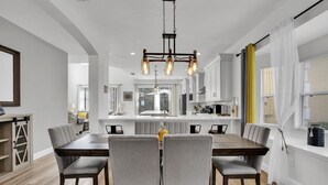 Ein moderner Essbereich mit Holztisch und grau gepolsterten Stühlen, einer Hängeleuchte sowie Blick auf die Küche und den Essraum durch eine Glastrennwand.