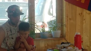 A man and a child sitting on a wooden bench inside a room with a window overlooking a parked vehicle. There is a small table with a red bottle and some food items on it. - Mountain View Guesthouse (Moron)