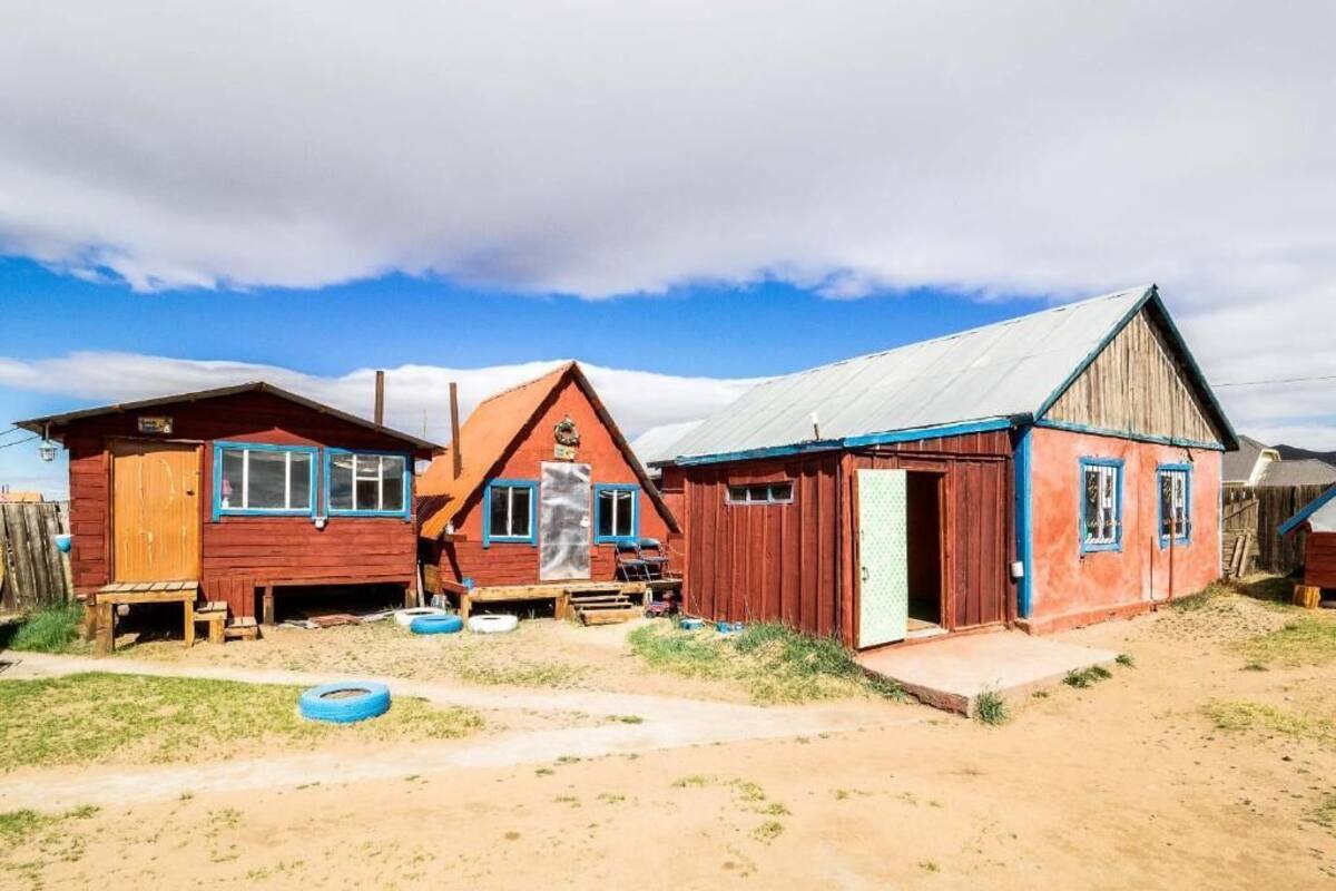 A rustic wooden house with a metal roof, a wooden porch, and a dirt yard with tires.