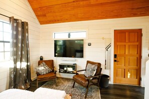 A cozy living room with a fireplace, two leather chairs with cushions, a flat-screen TV, and a wooden door. - Bluebonnet ↠ Cabin with view and Pro Coffee Setup, Pickleball (Wimberley)