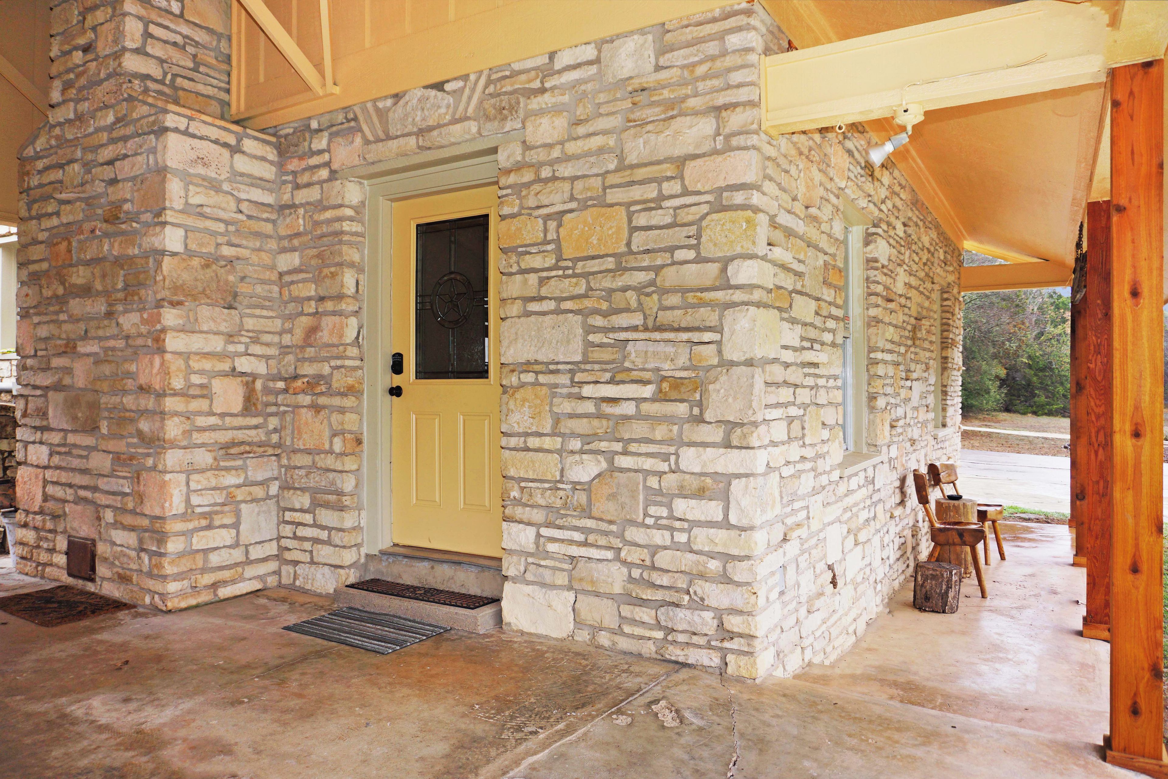 A stone building with a yellow door, a covered porch with wooden chairs, and a concrete floor.