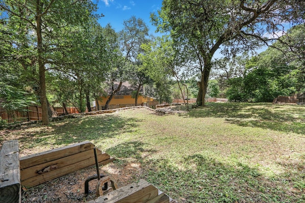 A fenced outdoor area with a wooden bench, trees, and a building in the background.