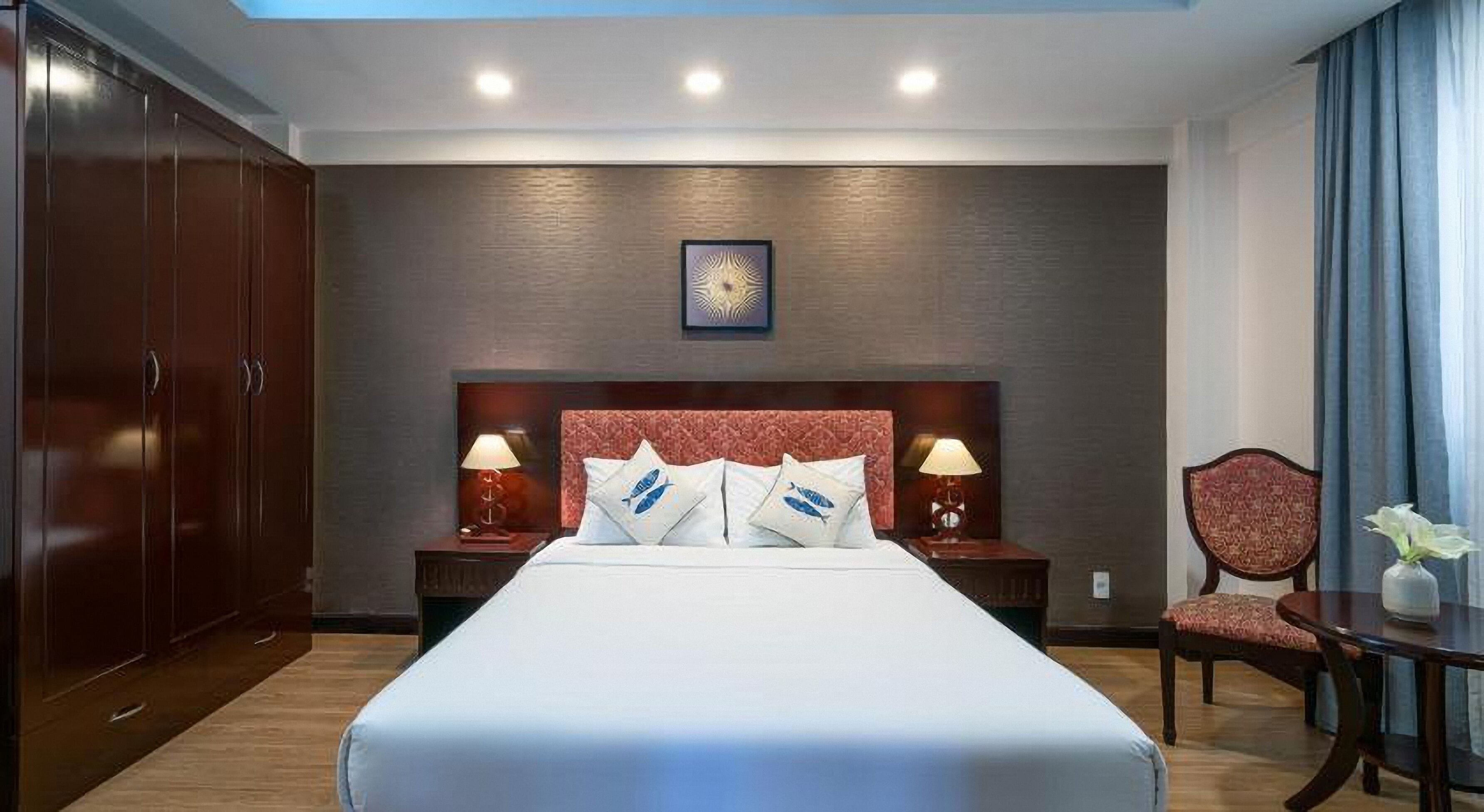 A hotel room with a large bed, two bedside tables with lamps, a wooden wardrobe, a red upholstered headboard, and a framed picture above the bed.