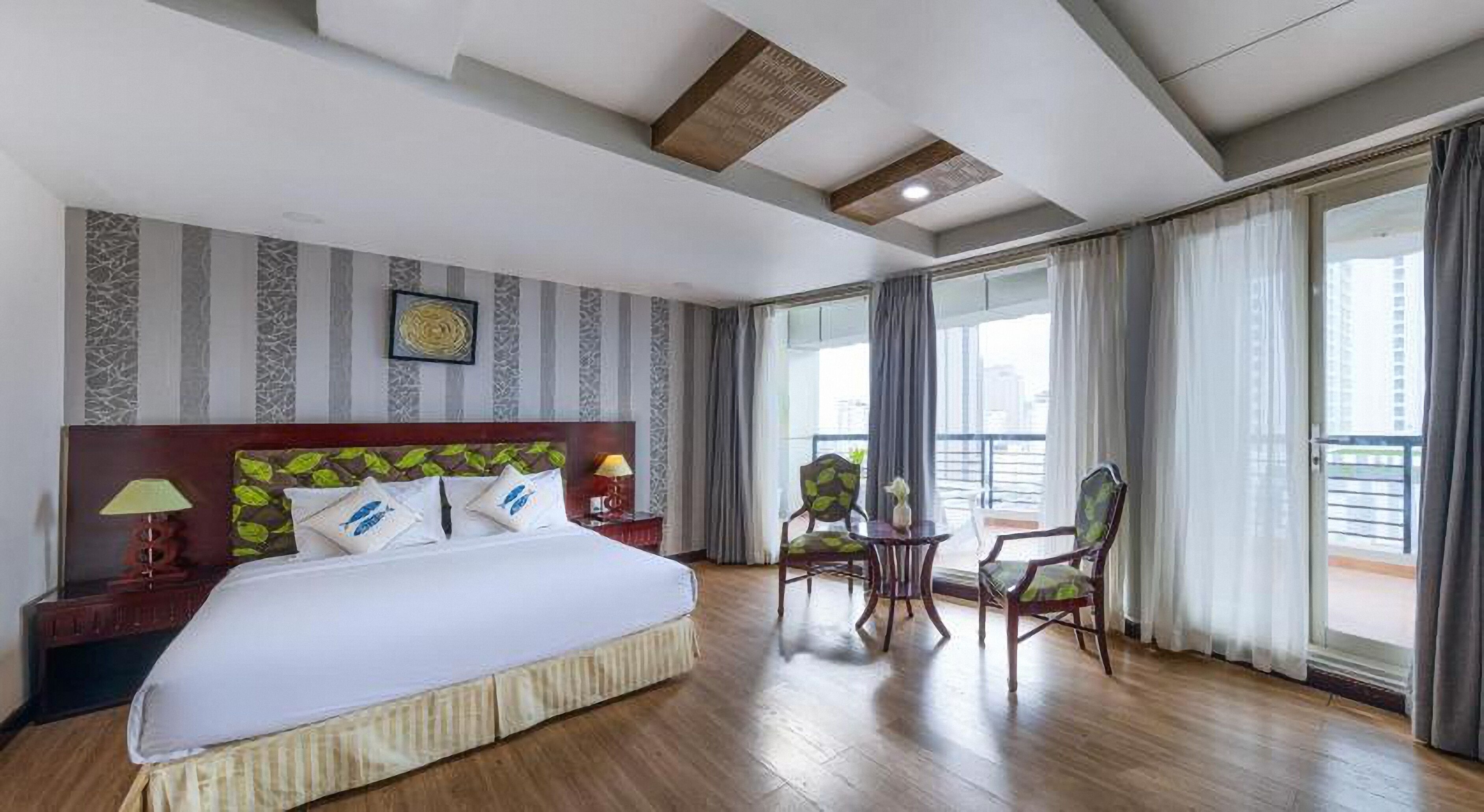 Habitación de hotel con una cama grande, una mesita con sillas, vistas de la ciudad a través de amplias ventanas y un balcón.