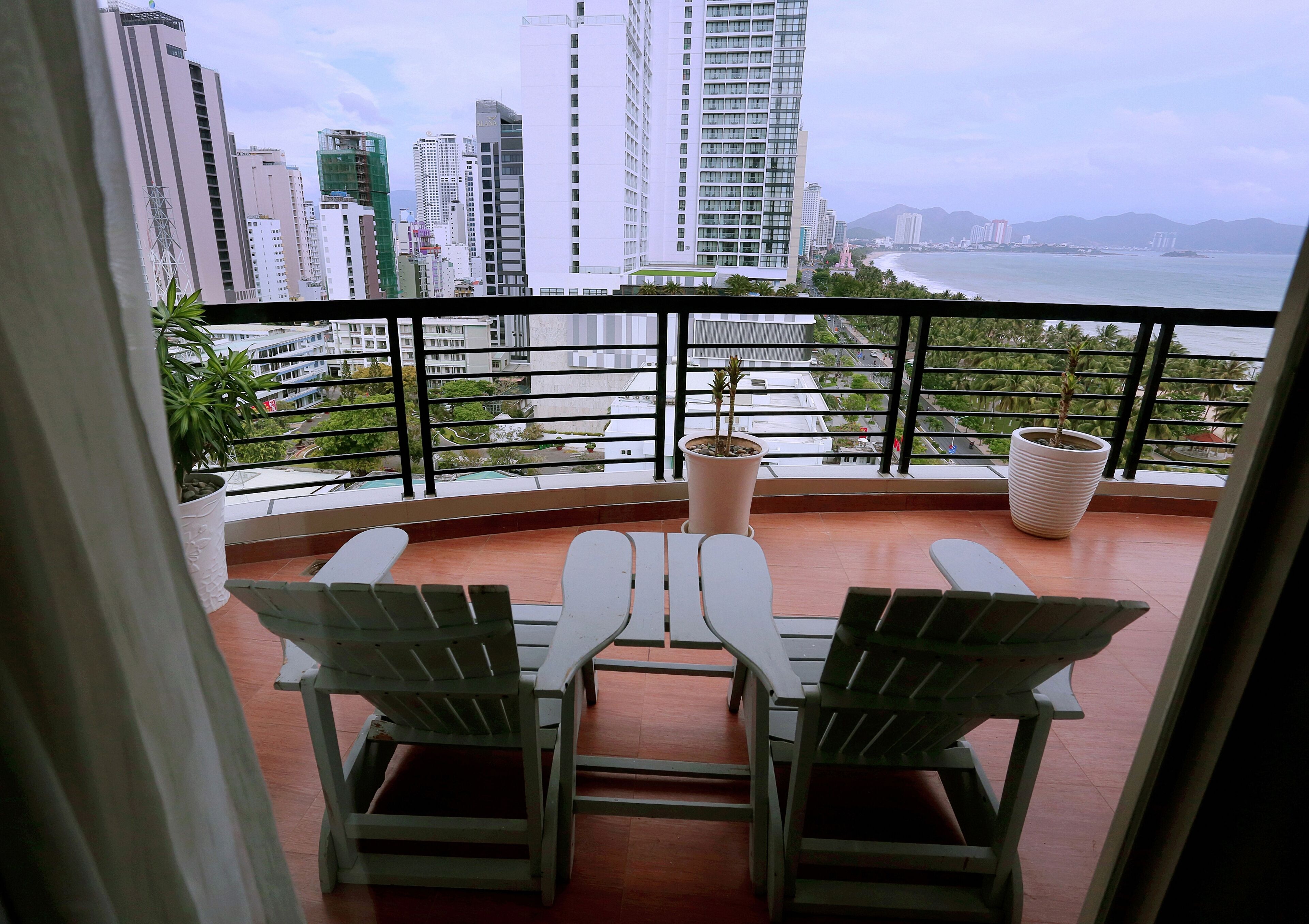 Un balcón con dos sillas blancas y una mesa, con vistas a una ciudad con edificios altos y una masa de agua.