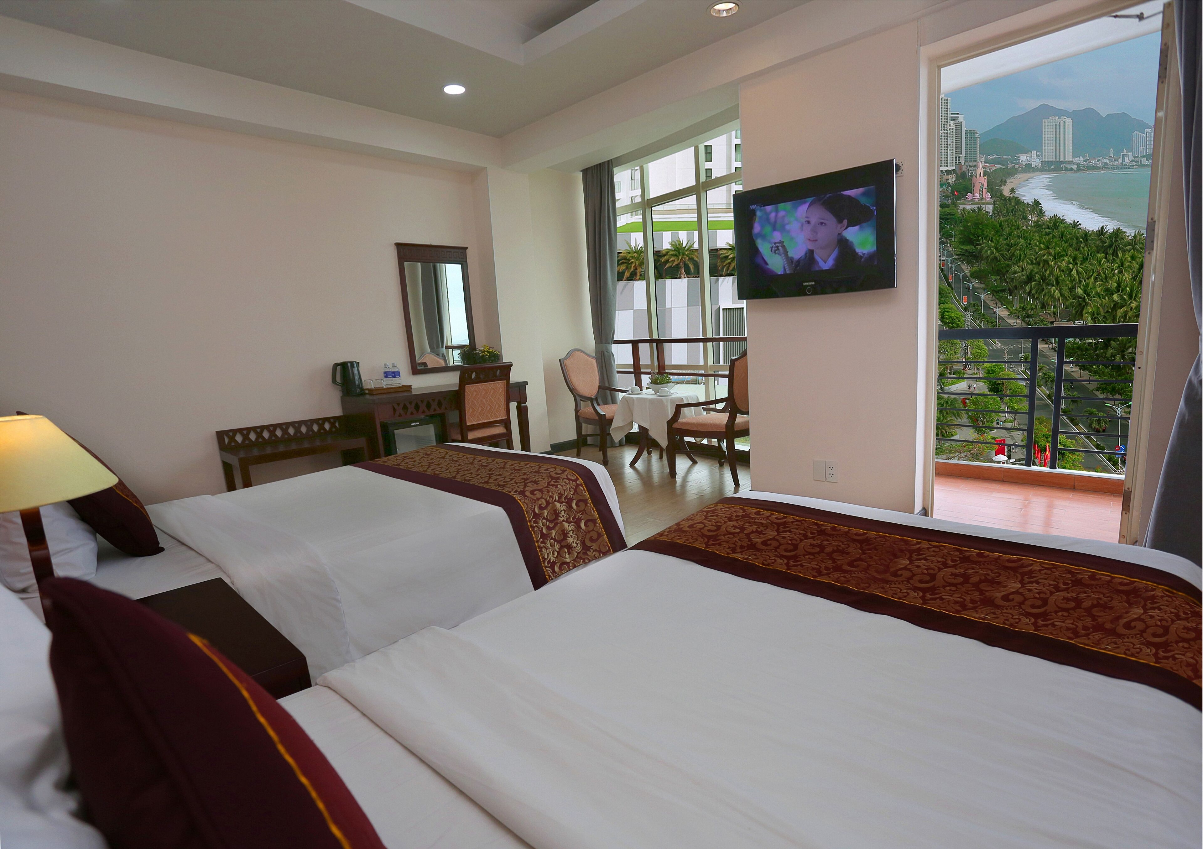 Habitación de hotel con dos camas, un escritorio, una silla, un televisor y un balcón con vistas a la ciudad y la playa.