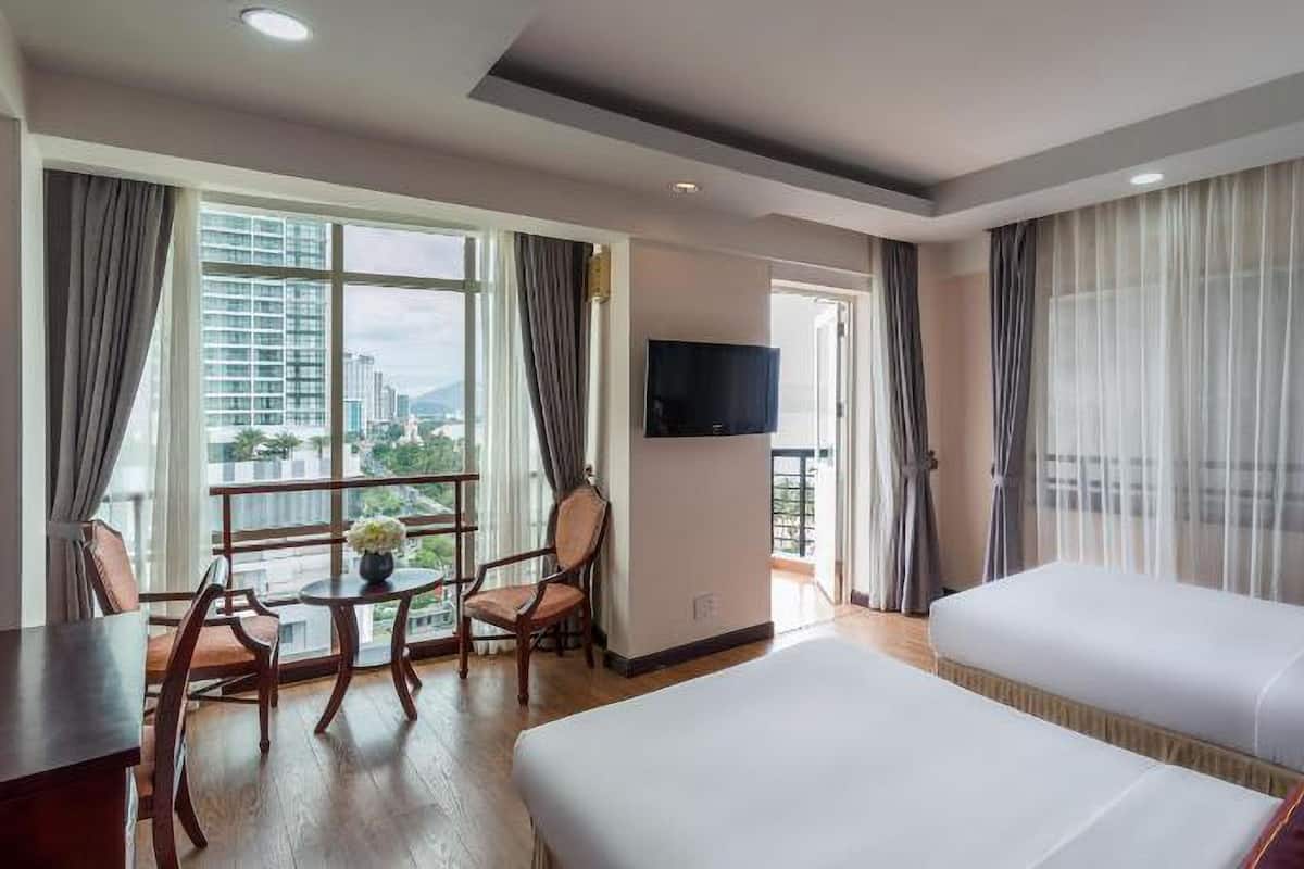 Habitación de hotel con dos camas, un escritorio con sillas, televisor y ventanas grandes con cortinas.