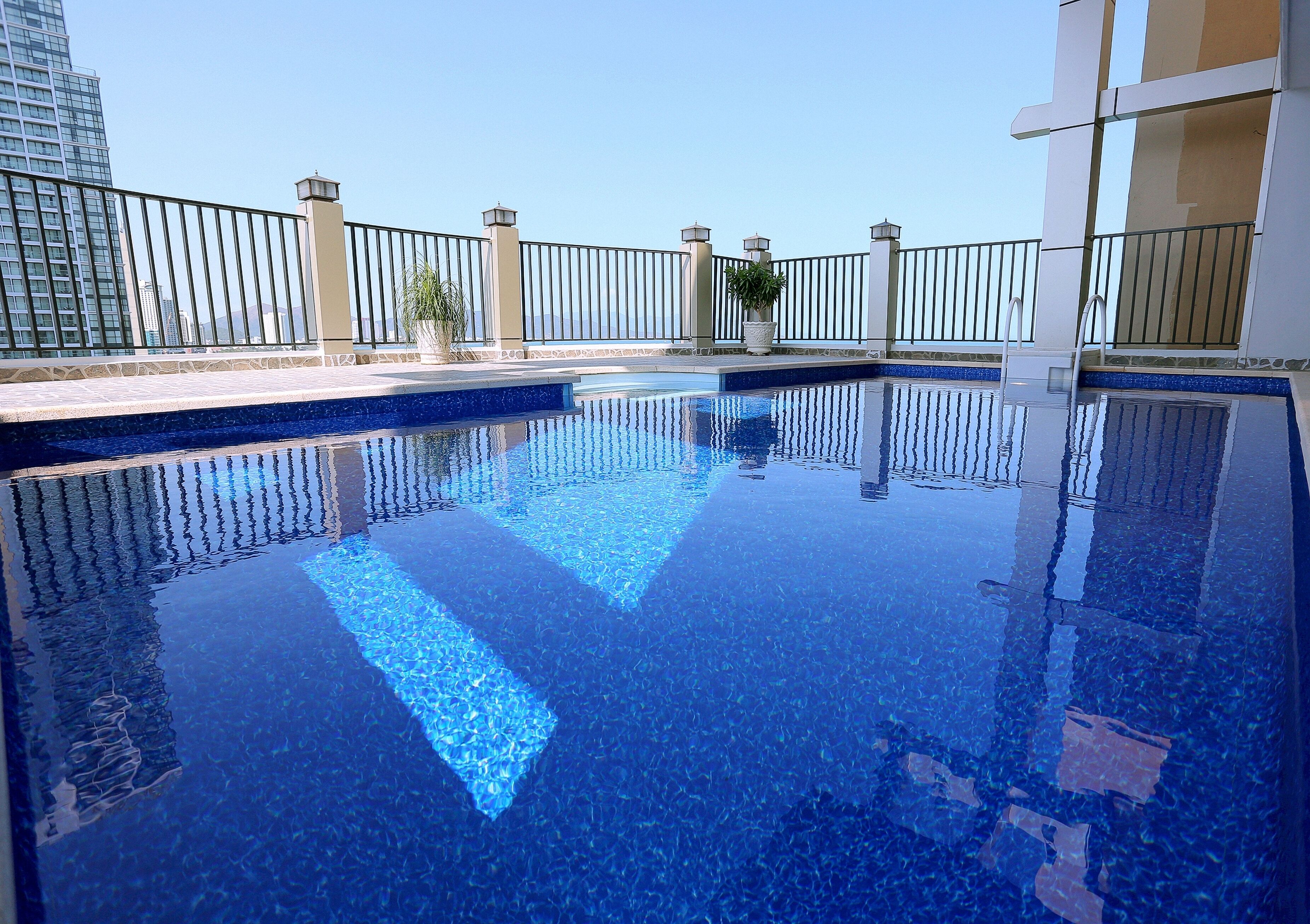 Une piscine aux eaux d’un bleu limpide, bordée de carreaux, entourée d’une clôture métallique et agrémentée de plantes en pot.
