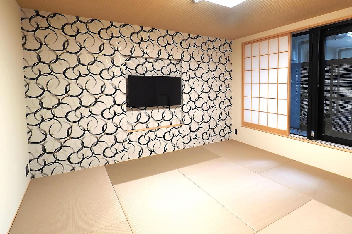 Una habitación con suelo de tatami, un televisor negro montado en una pared estampada y una puerta corredera tipo shoji.