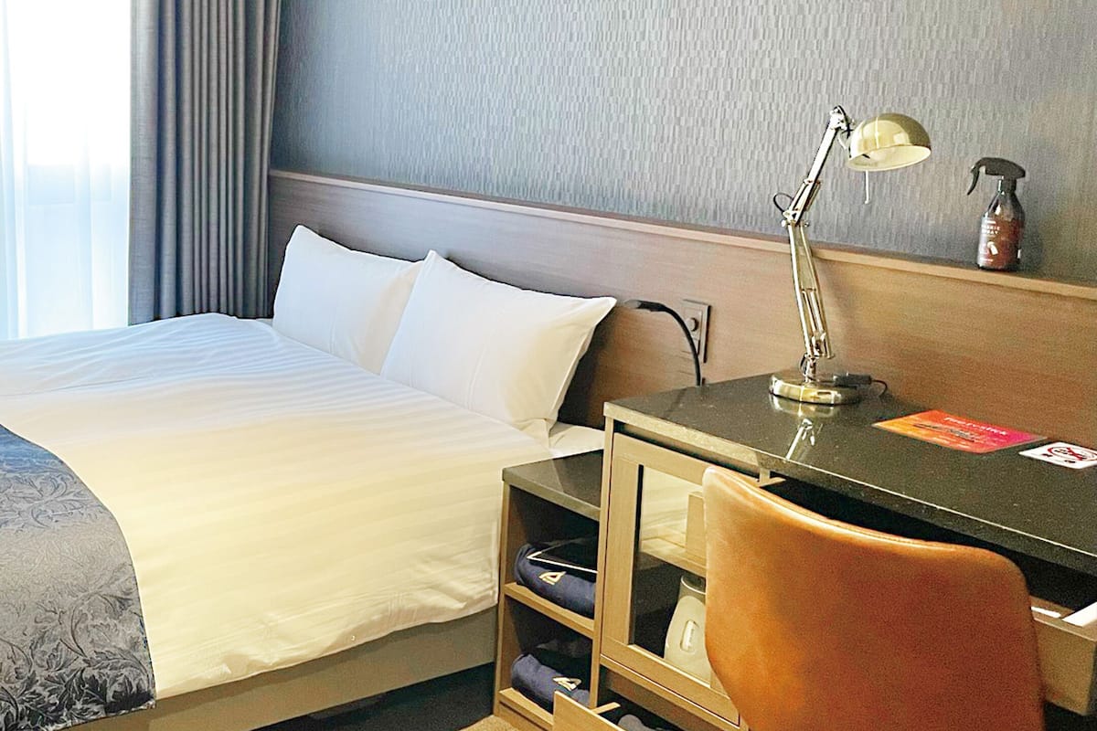 Habitación de hotel con cama, escritorio, silla, lámpara y estante pequeño con una botella y un libro.