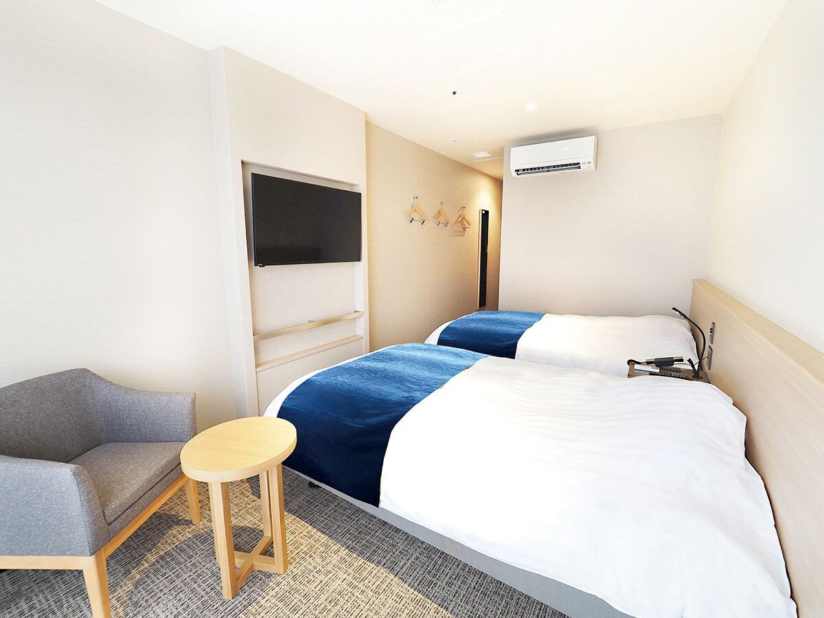 A hotel room with a bed, a chair, a small table, a TV, and an air conditioning unit.