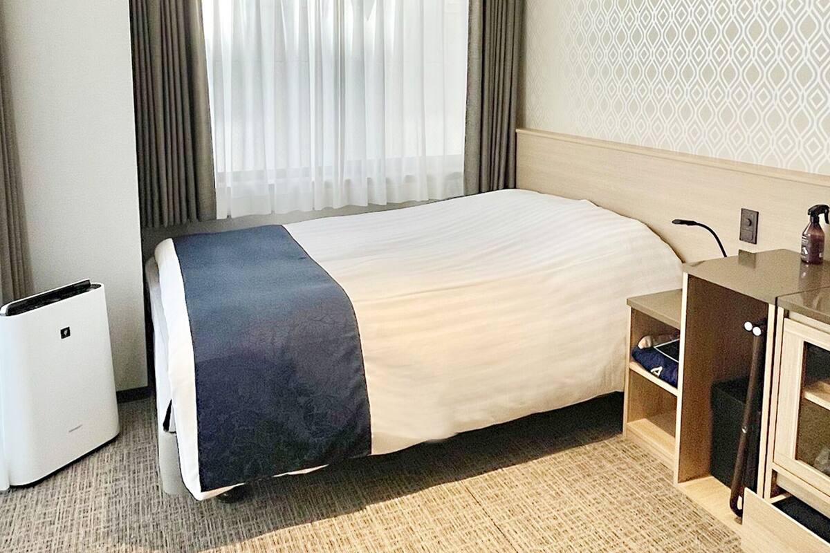 Habitación de hotel con cama, escritorio, silla, cubo de basura y ventana con cortinas.
